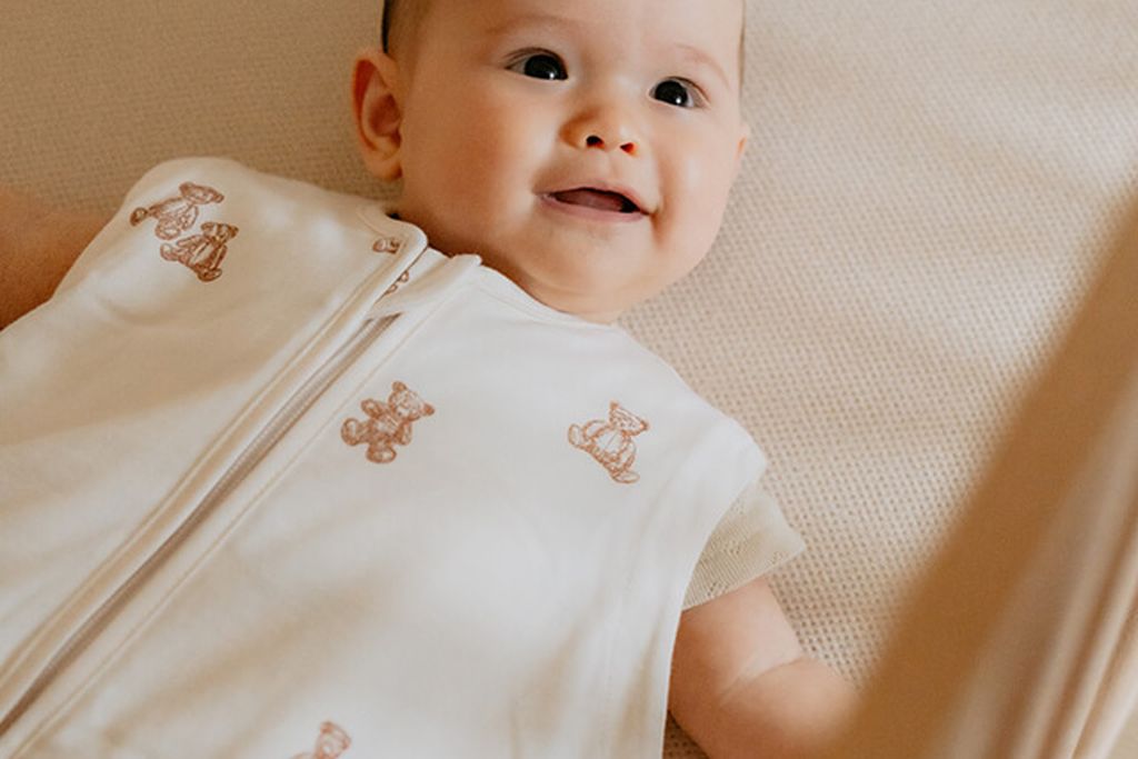 Een glimlachende baby ligt op een zacht oppervlak, draagt een Puckababy slaapzak met Teddy Bears-print en kijkt omhoog.