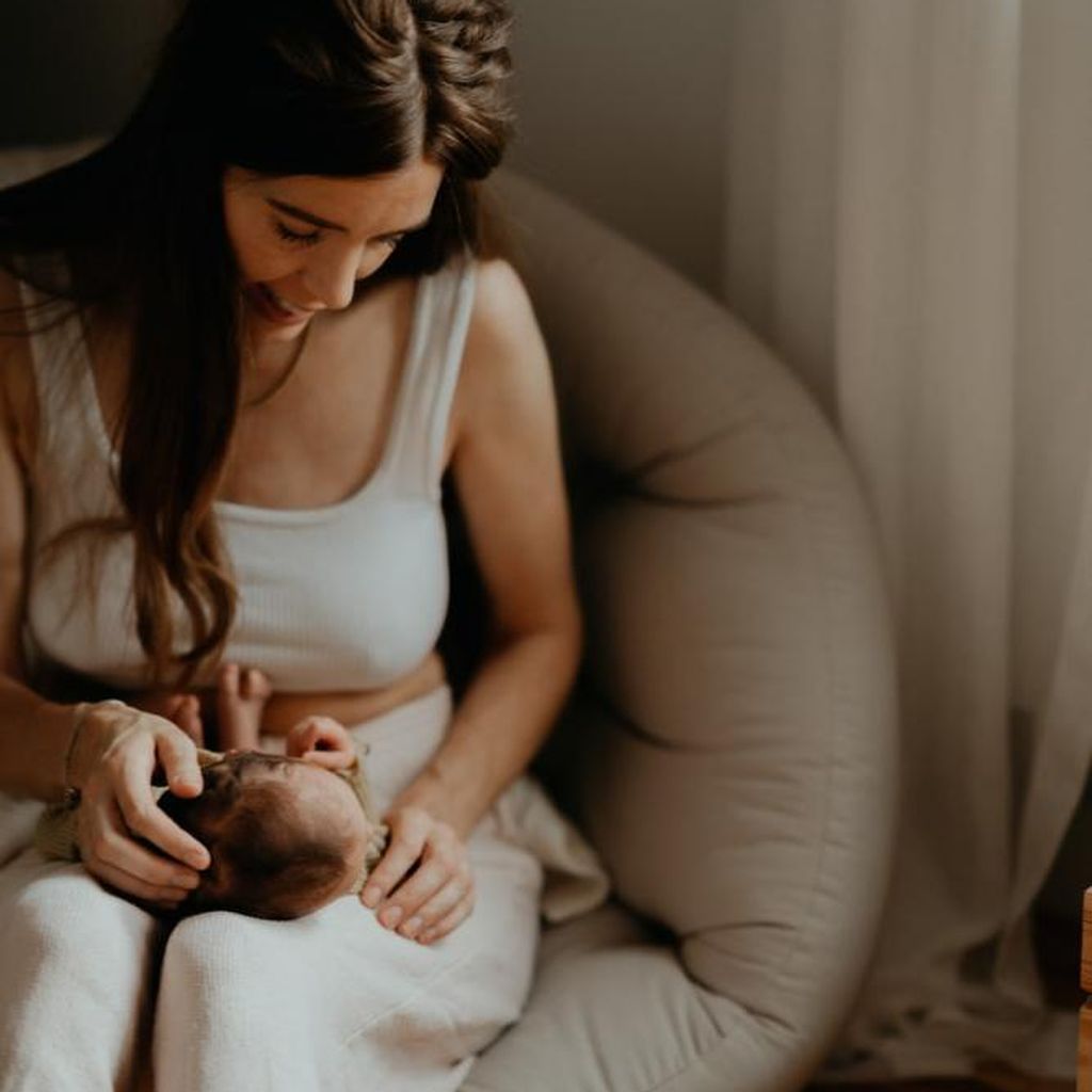 Eine Frau sitzt in einem gemütlichen Sessel, hält sanft ein Baby im Arm und schaut es an.