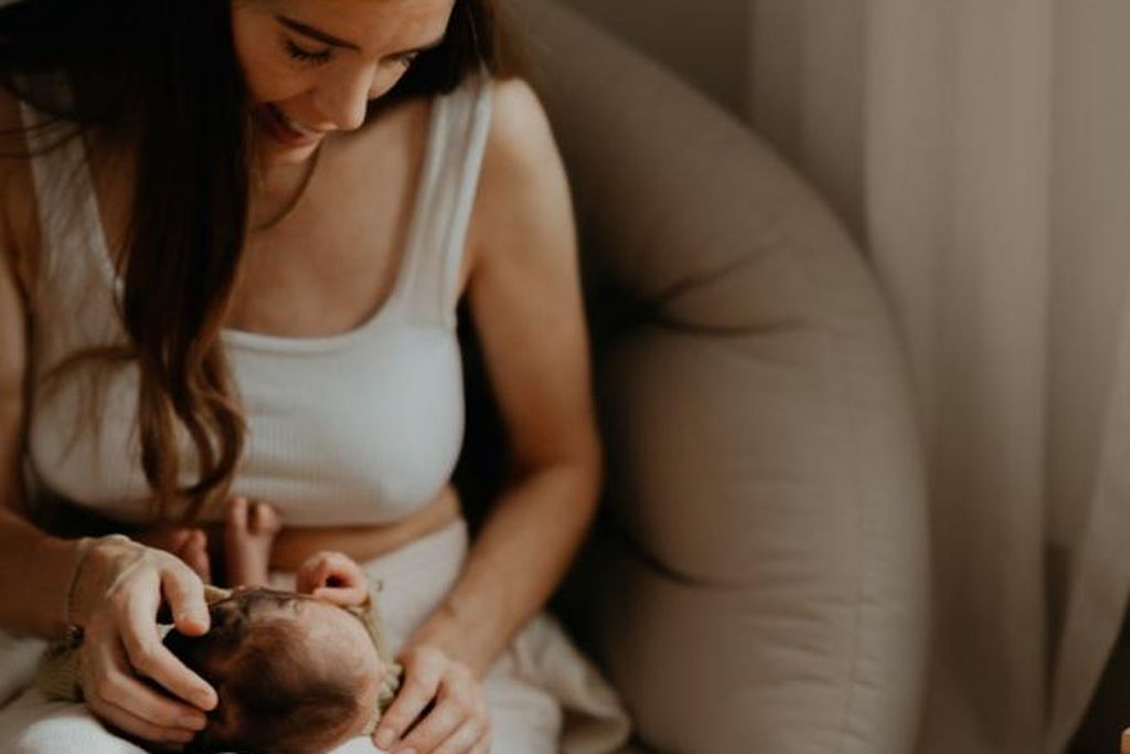 A woman sits in a cozy chair, gently holding and looking at a baby wrapped