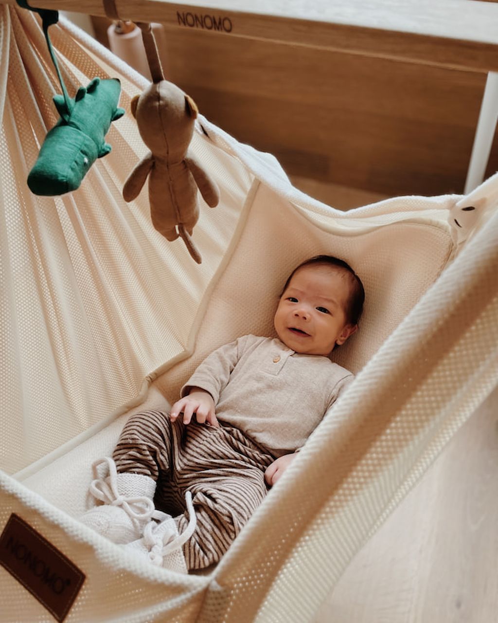 Ein Baby liegt in einer cremefarbenen Hängematte und lächelt. Plüschtiere hängen darüber, und die Hängematte