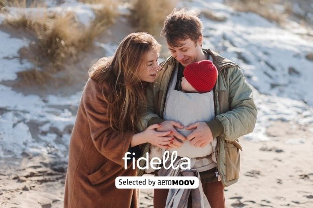 Een koppel in winterkleding draagt een baby in een draagzak op een besneeuwd strand. De vrouw staat links en de man rechts. Tekst: “Fidella geselecteerd door AeroMoov”.