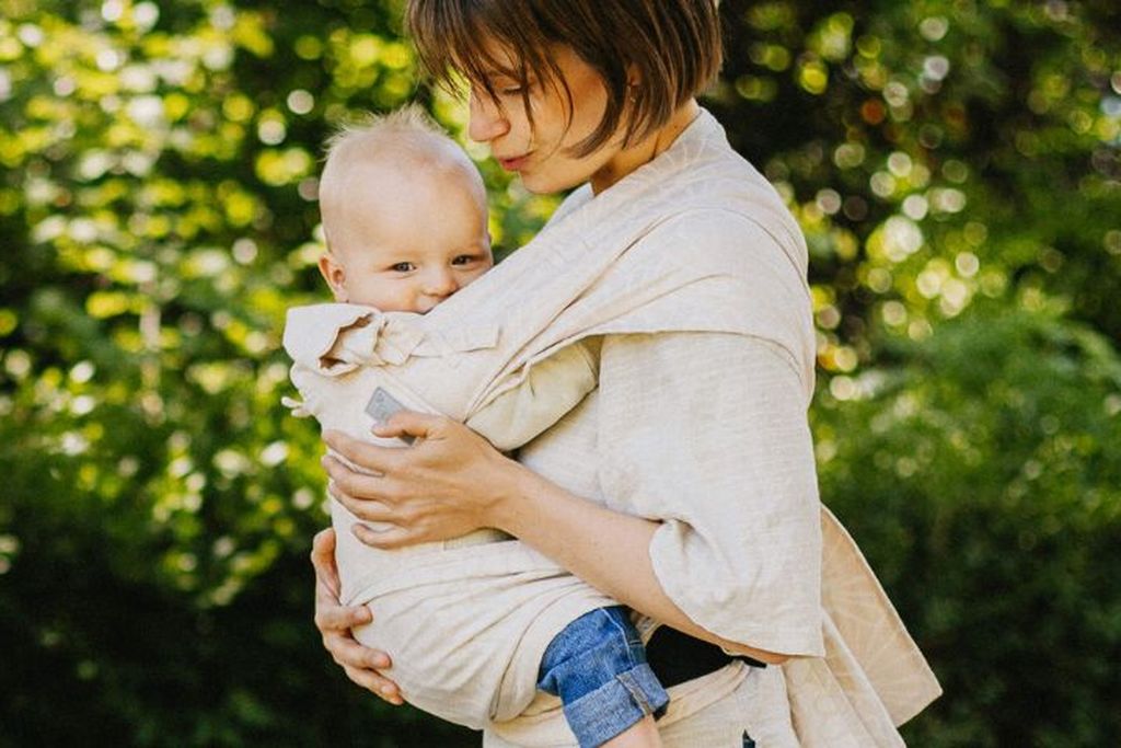 Une femme tient un bébé dans un FlyClick beige, à l’extérieur devant un fond flou de feuillage vert.
