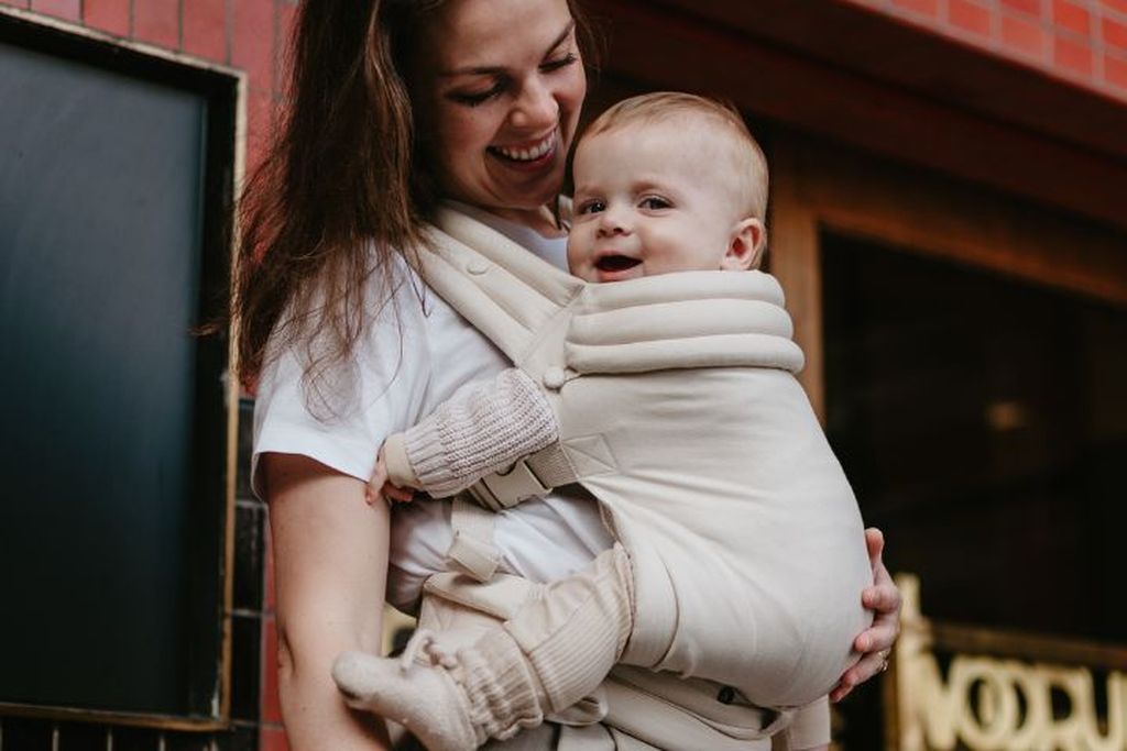 Smiling woman holding a happy baby in a beige carrier, standing outside a building with a red brick facade.