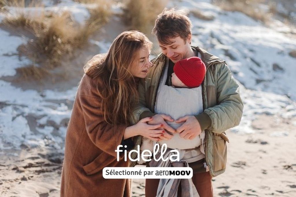Un couple en vêtements d’hiver porte un bébé dans un porte-bébé sur une plage enneigée. La femme est à gauche et l’homme à droite. Texte : « Fidella sélectionné par AeroMoov ».