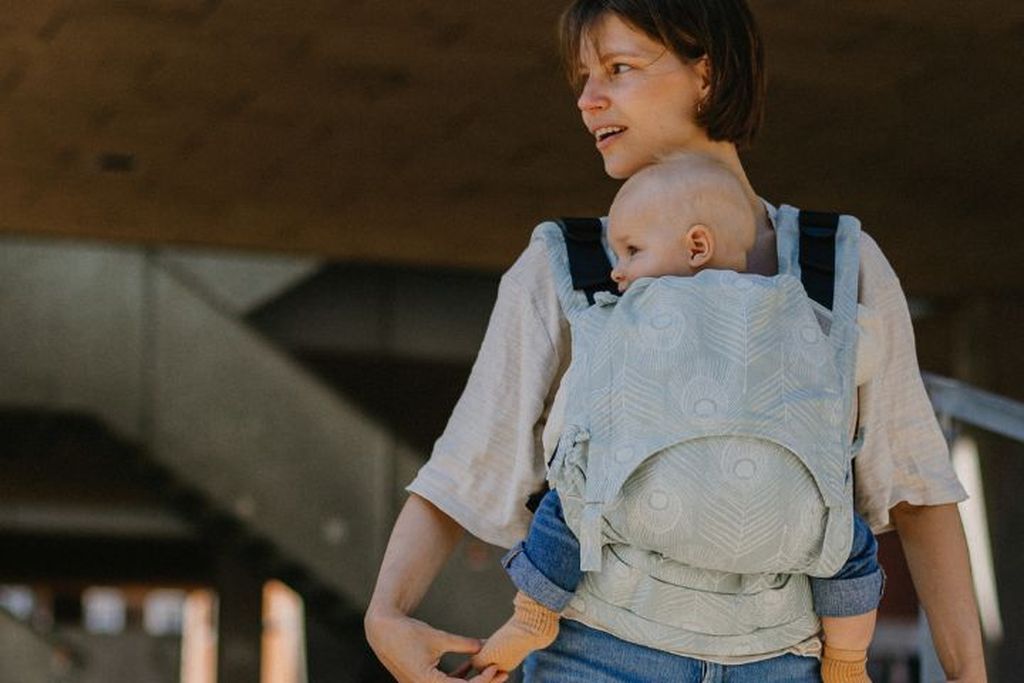 Une femme porte un bébé dans un porte-bébé à motifs, tous deux regardant sur le côté. Ils sont dehors sous une structure ombragée.
