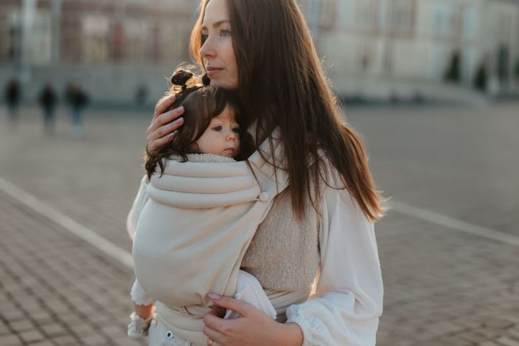 Woman holding a baby in a carrier, standing outdoors. Both are looking in the same direction with a serene expression.