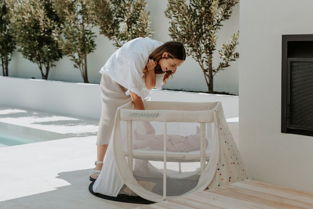 Vrouw die voor een baby zorgt in een AeroMoov Instant Travel Cot bij een zwembad, met bomen en een witte muur op de achtergrond