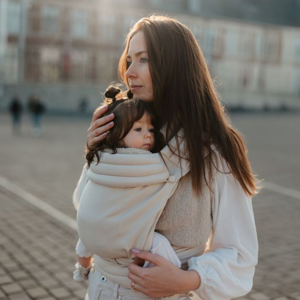 A woman holding a baby in a beige carrier outdoors, looking into the distance with a soft expression. The background shows a blurred building.