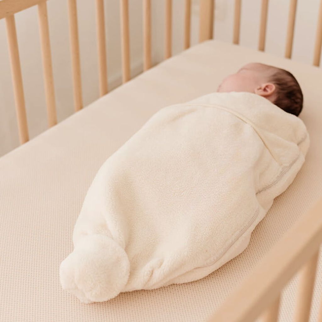 Een baby is gewikkeld in een zachte, crèmekleurige deken en slaapt vredig in een houten wieg met een AeroSleep-matras.