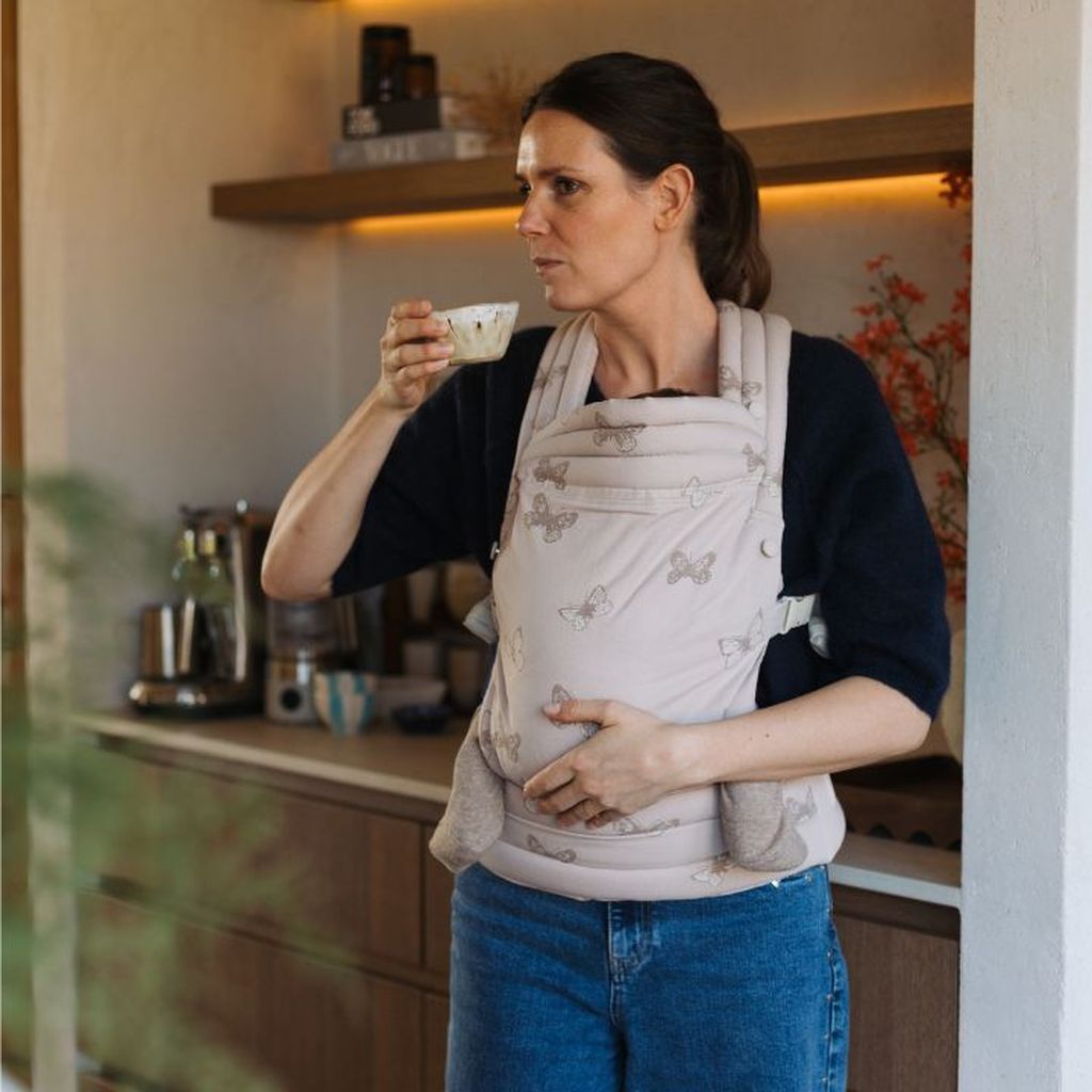 Woman holding a cup of coffee, wearing a baby carrier with a butterfly pattern, standing in a kitchen.