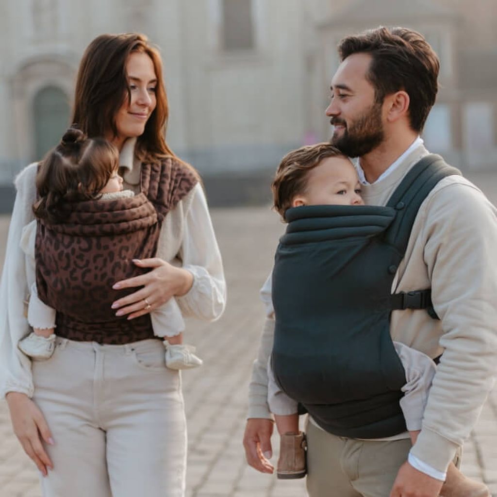A couple smiling at each other, each carrying a baby in front-facing carriers, standing outdoors on a paved area.