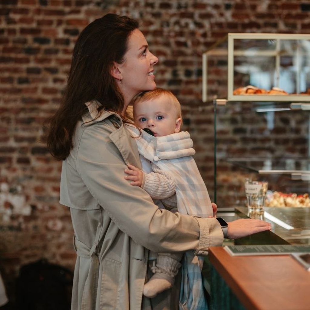 A woman in a trench coat holds a baby in a café, standing by a pastry display with a brick wall background.