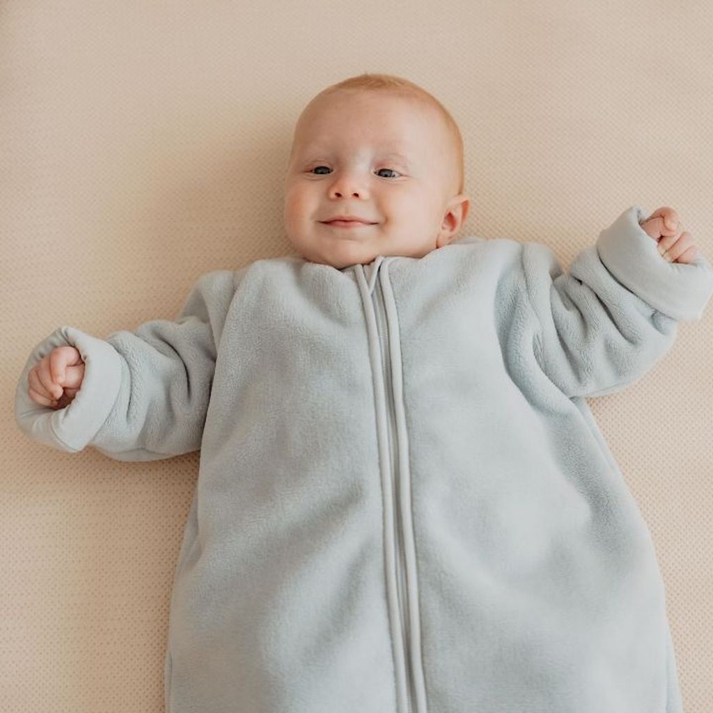 Smiling baby lying on a beige surface, wearing a light blue onesie with arms slightly raised.