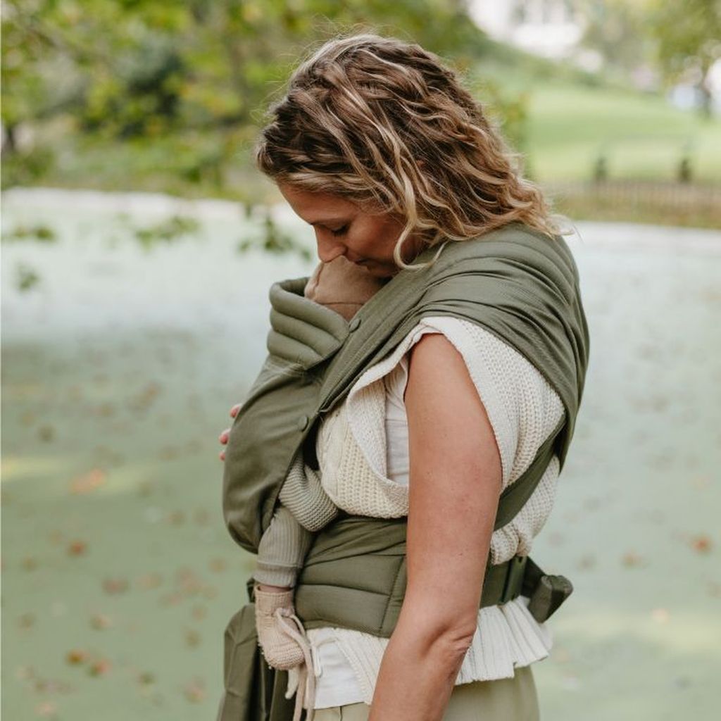 Woman gently embraces a baby in a green wrap carrier outdoors, surrounded by lush greenery.