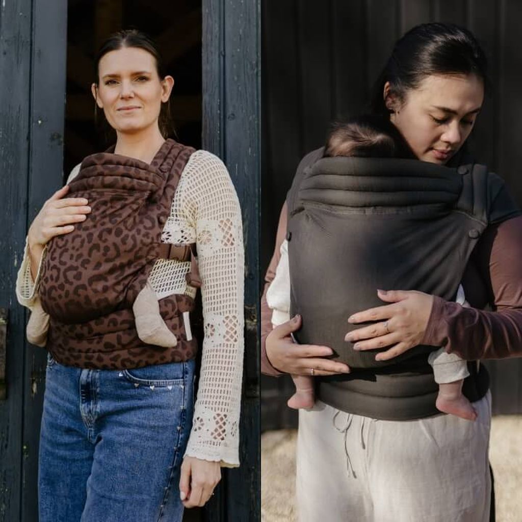 Two women each carrying a baby in soft carriers. One carrier has a leopard print, and the other is plain.