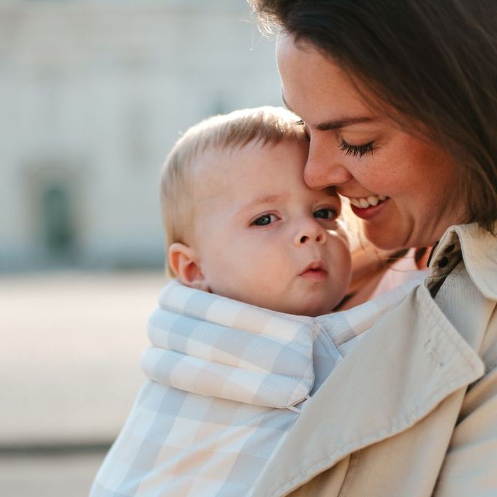 A woman lovingly embraces a baby wrapped in a checkered blanket, both smiling softly, creating a tender moment.