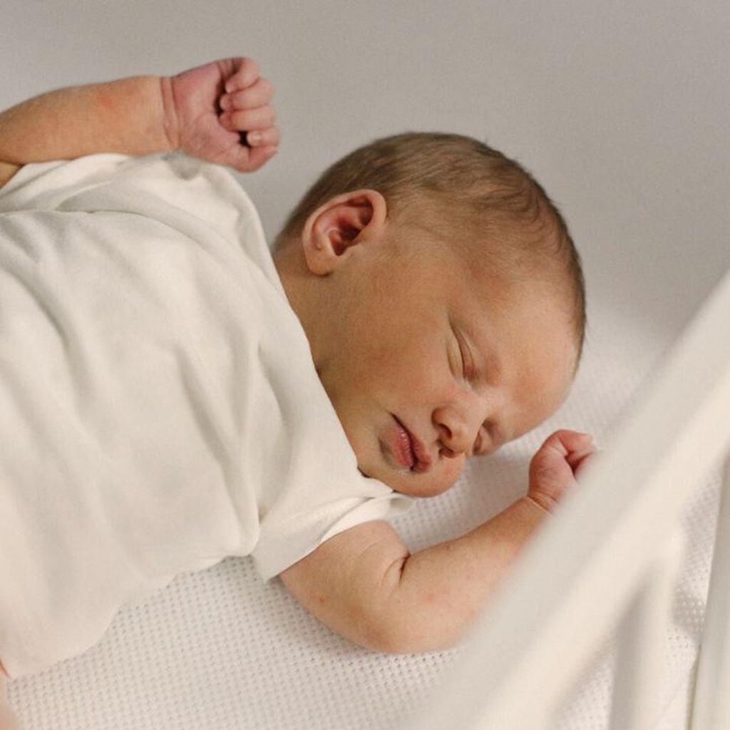 Newborn baby in a white outfit sleeping peacefully on a white mattress, with one arm raised and the other resting by their side.