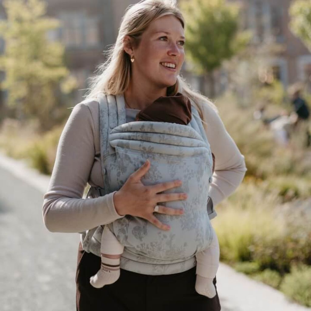 Woman walking outdoors, carrying a baby in a patterned carrier. She is smiling, with trees and buildings in the background.