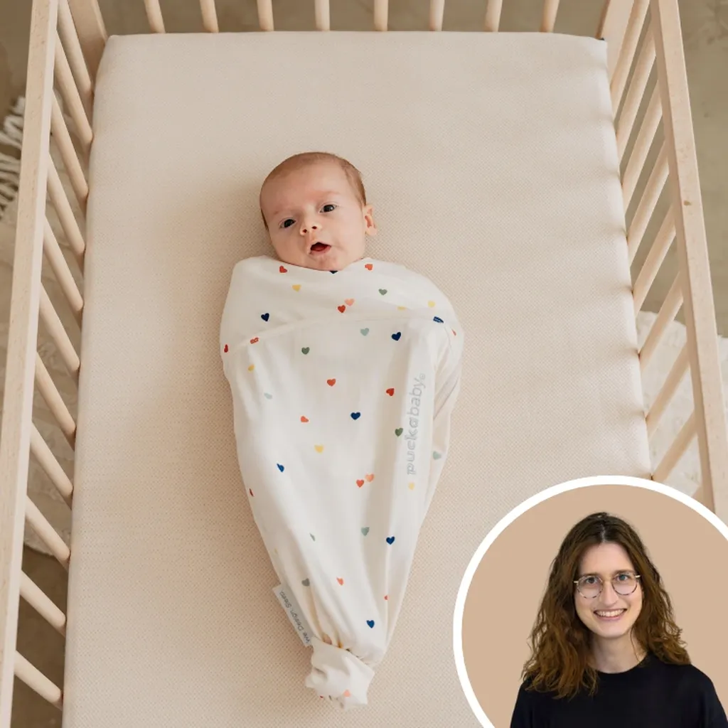 Baby ligt op een AeroSleep-matras in een inbakerslaapzak met hartjesmotief en geeuwt.