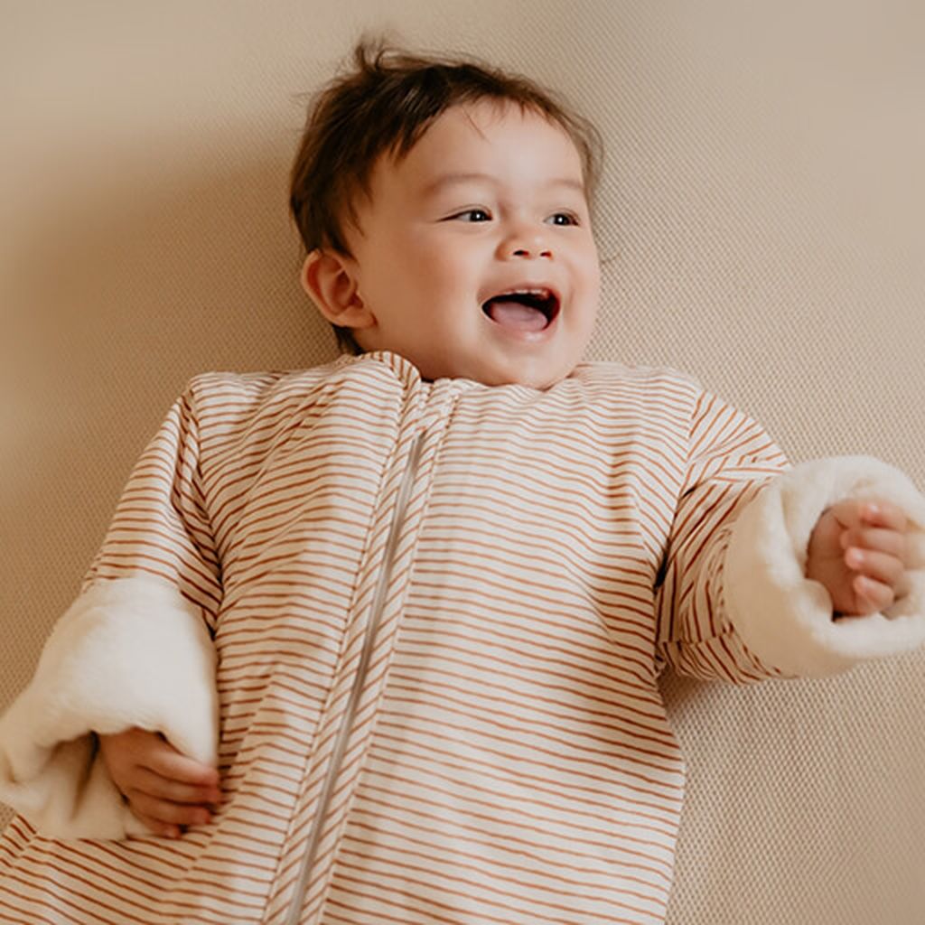 Een glimlachende peuter in een Puckababy Bag 4 Seasons met Hazel Stripes-print.