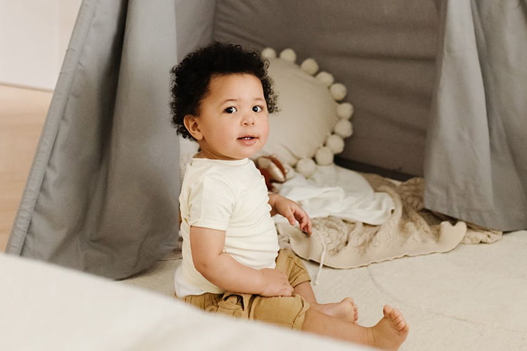 A toddler sits inside a gray play tent, surrounded by pillows and blankets