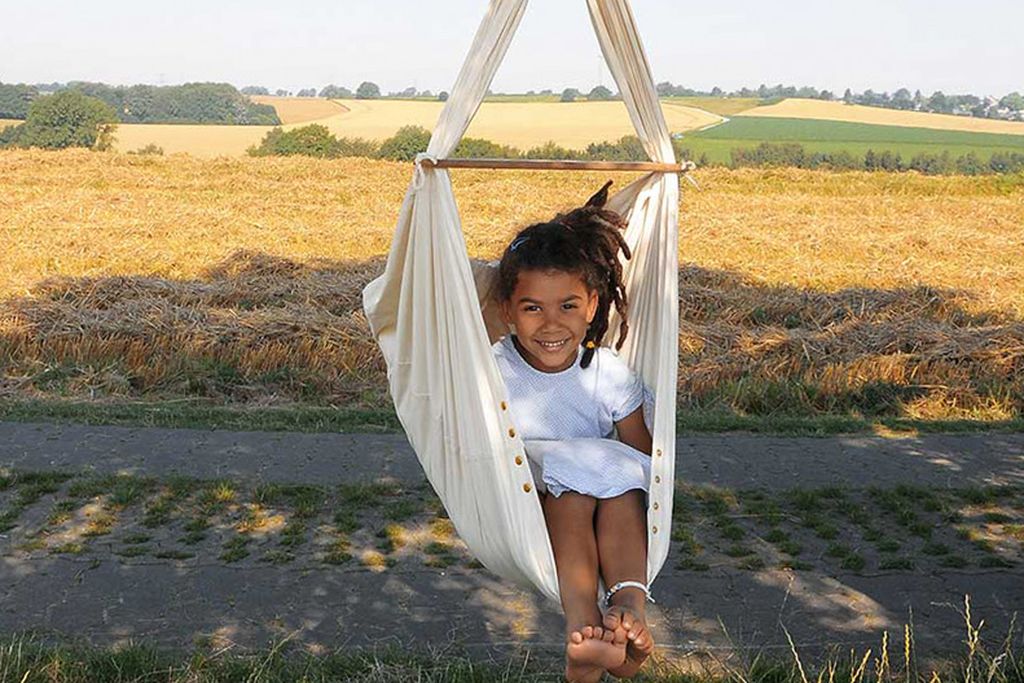 Ein lächelndes Mädchen sitzt in einer Kleinkindhängematte im Freien, mit einem Feld im Hintergrund.