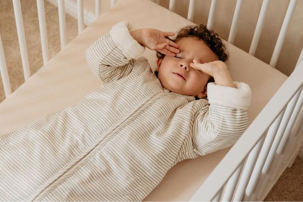 A toddler in a striped Bag 4 Seasons rubs their eyes while lying in a crib
