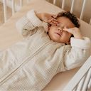 A toddler in a striped Bag 4 Seasons rubs their eyes while lying in a crib