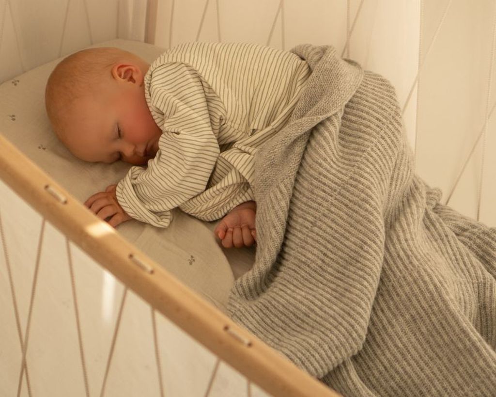 A baby sleeps peacefully in a crib, wrapped in a gray knitted blanket, wearing a striped bag 4 seasons