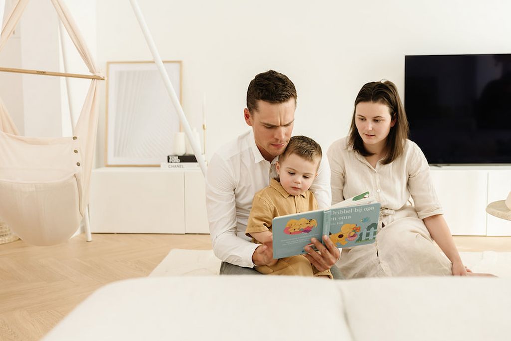Eine dreiköpfige Familie sitzt auf dem Boden in einem hellen Wohnzimmer, wobei der Vater seinem kleinen Kind aus einem Buch vorliest