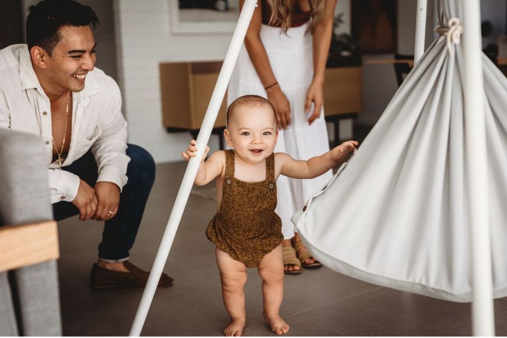 Ein lächelndes Baby steht neben einer Federwiege und hält ein Bein des Tipi Gestells fest