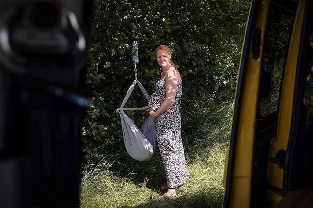 Eine schwangere Frau steht draußen in der Natur vor einer grauen Baby Federwiege