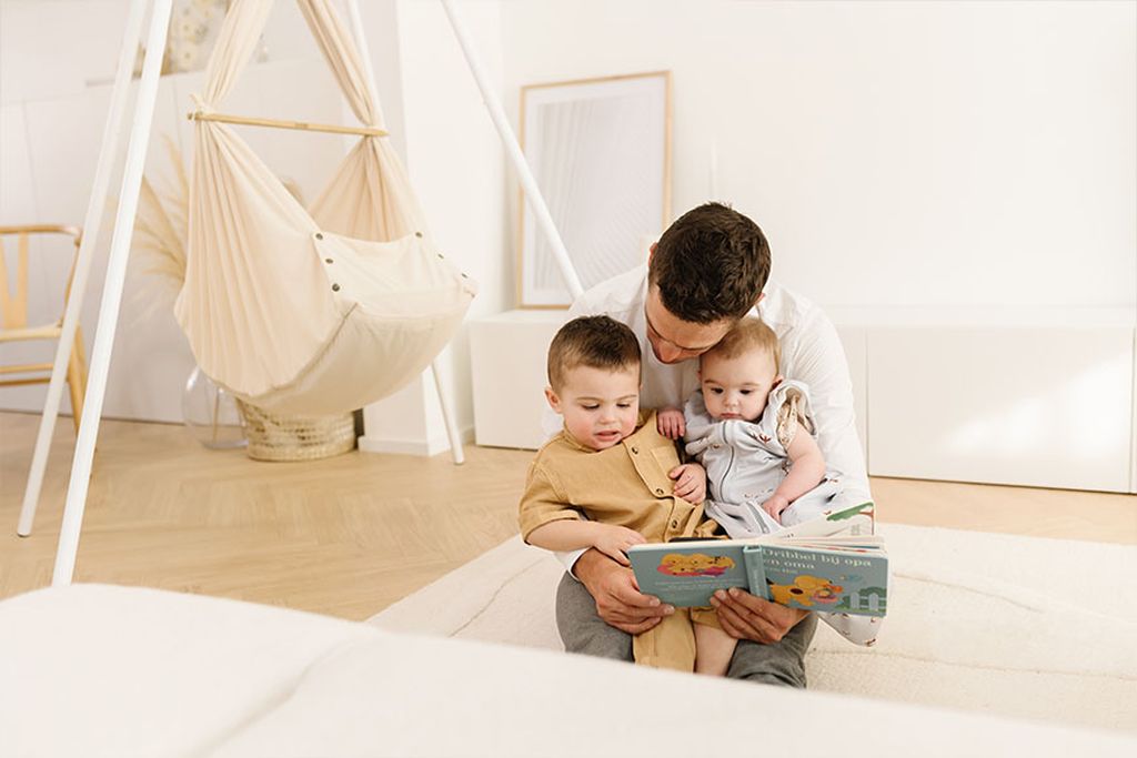 EinVater hat zwei Kinder auf dem Schoß und liest ihnen ein Buch vor.