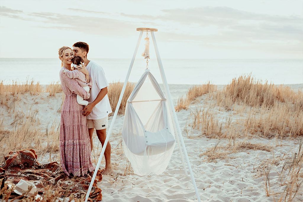 Famile am Strand. Im Vordergrund ist eine NONOMO Federwiege an einem Tipi Gestell befestigt