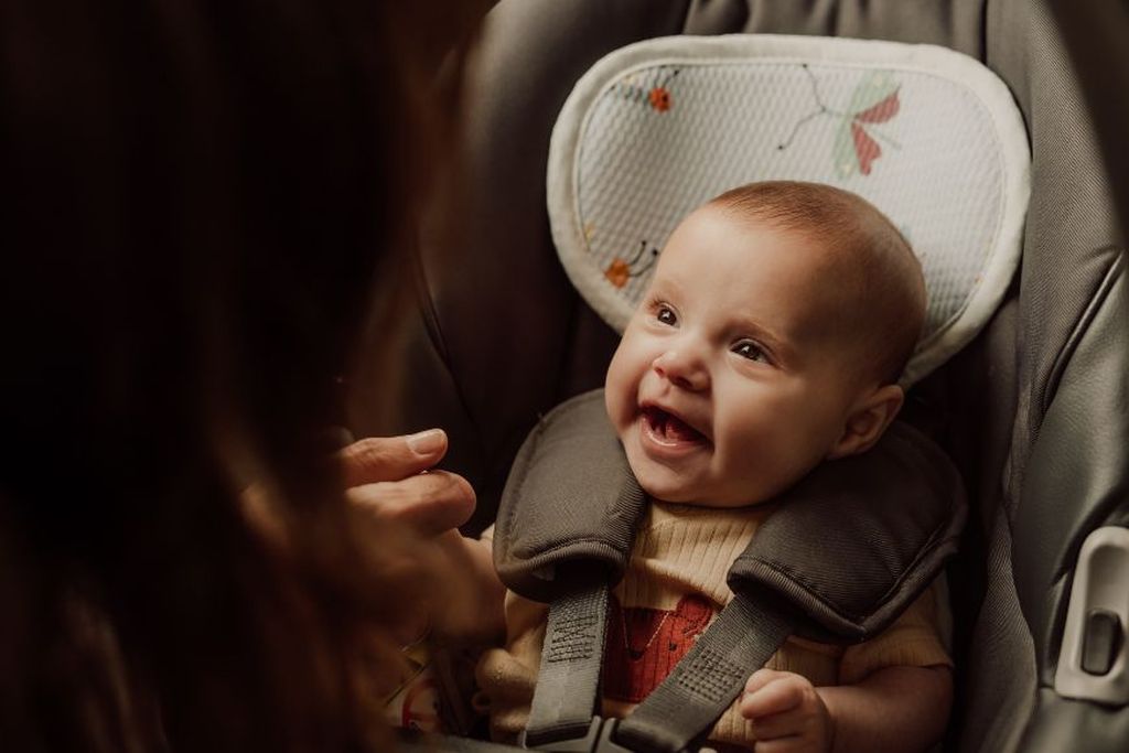 Een lachende baby in een autostoeltje kijkt omhoog naar een persoon. Zittend in een Air Layer
