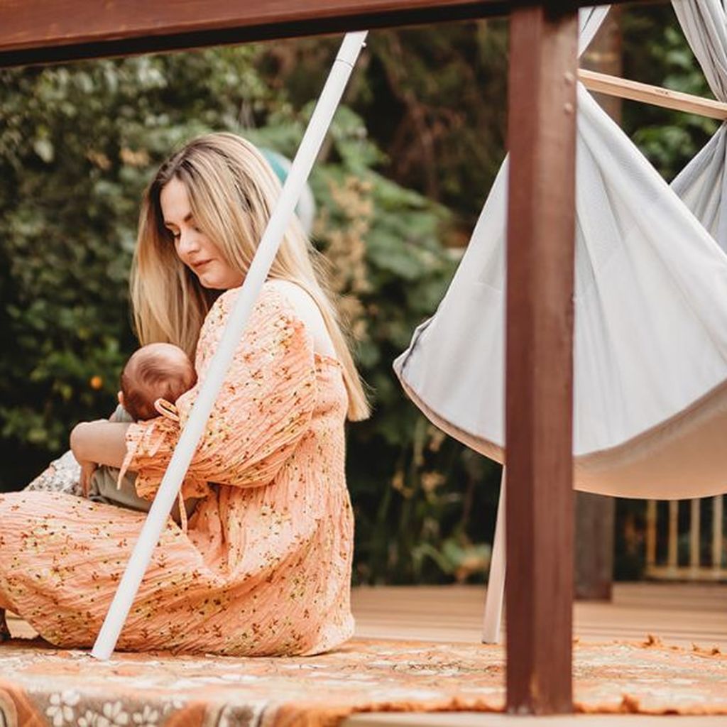 Eine Frau in einem geblümten Kleid sitzt auf einer Holzterrasse und wiegt ein Baby. Im Hintergrund ist eine Federwiege zu sehen.