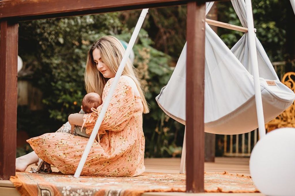 Eine Frau in einem geblümten Kleid sitzt auf einer Holzterrasse und wiegt ein Baby in den Armen. Im Hintergrund ist eine Hängematte zu sehen.