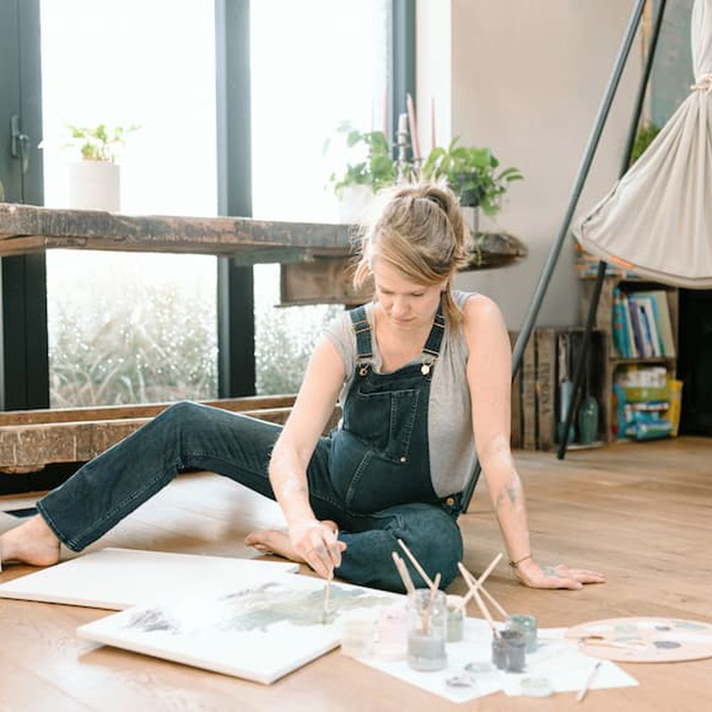 A pregnant woman sitting on the floor painting on a canvas in the background a hammock is hanging