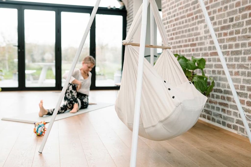 Im Mittelpunkt steht die Babyhängematte in einem Tipi-Ständer, und im Hintergrund praktiziert eine Frau Yoga.