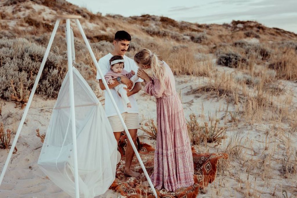 Eine Familie mit einem Baby steht an einem Sandstrand in der Nähe eines weißen Tipi-Ständers, an dem eine Federwiege befestigt ist.