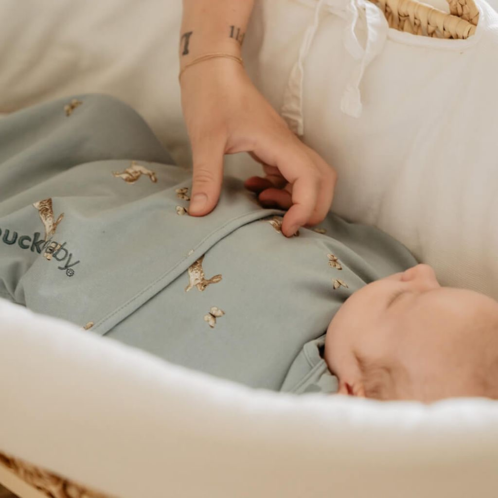 Baby gewikkeld in een Piep inbakerslaapzak met Bunnies-print, liggend in een mand. Een volwassen hand past voorzichtig de inbakerdoek aan.