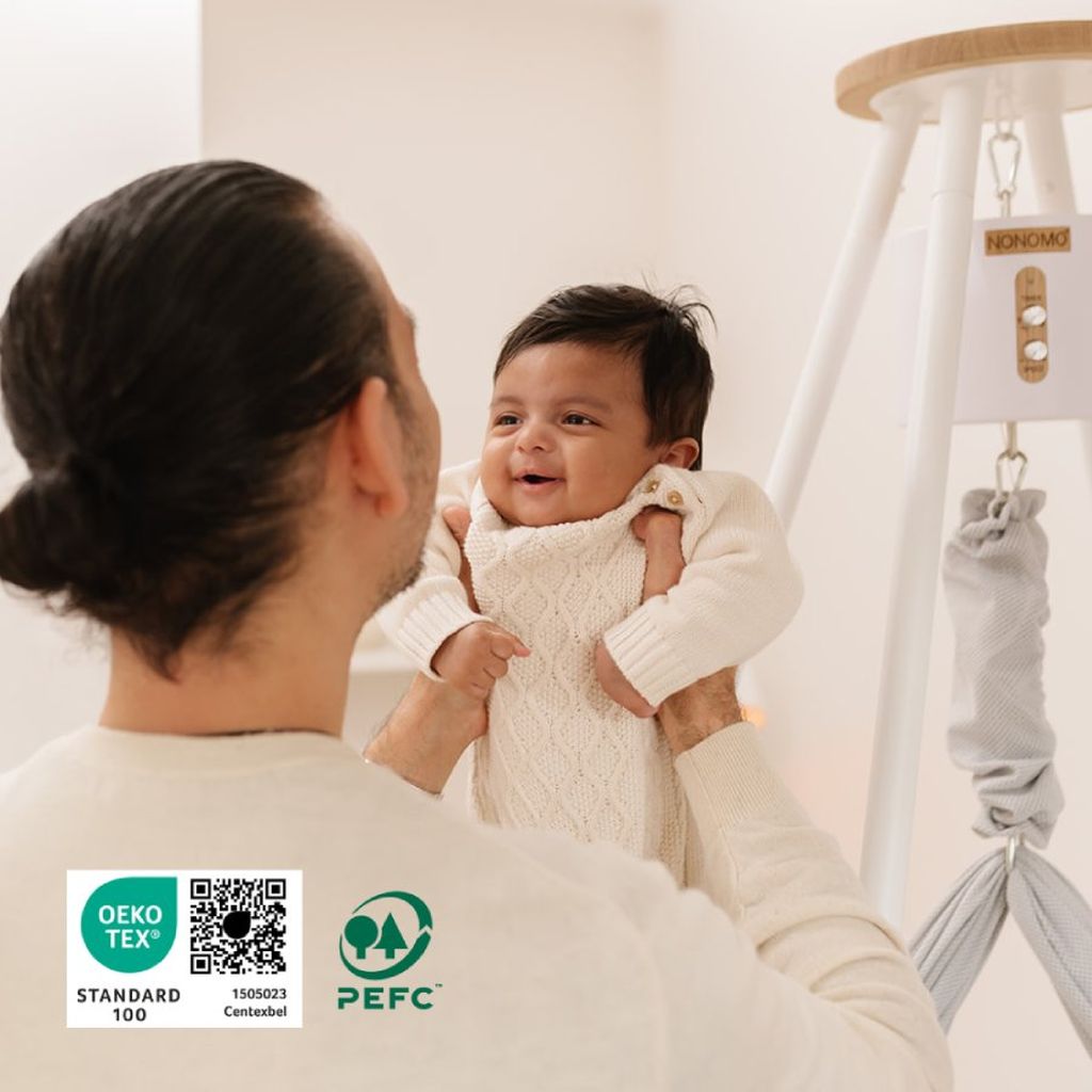 A person holding a smiling baby with a hammock in the background. Certification logos for OEKO-TEX and PEFC visible in the corner