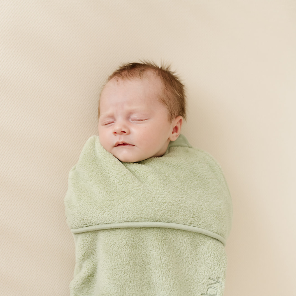 Een slapende baby gewikkeld in een Puckababy Piep Teddy Olive, liggend op een lichtbeige achtergrond.