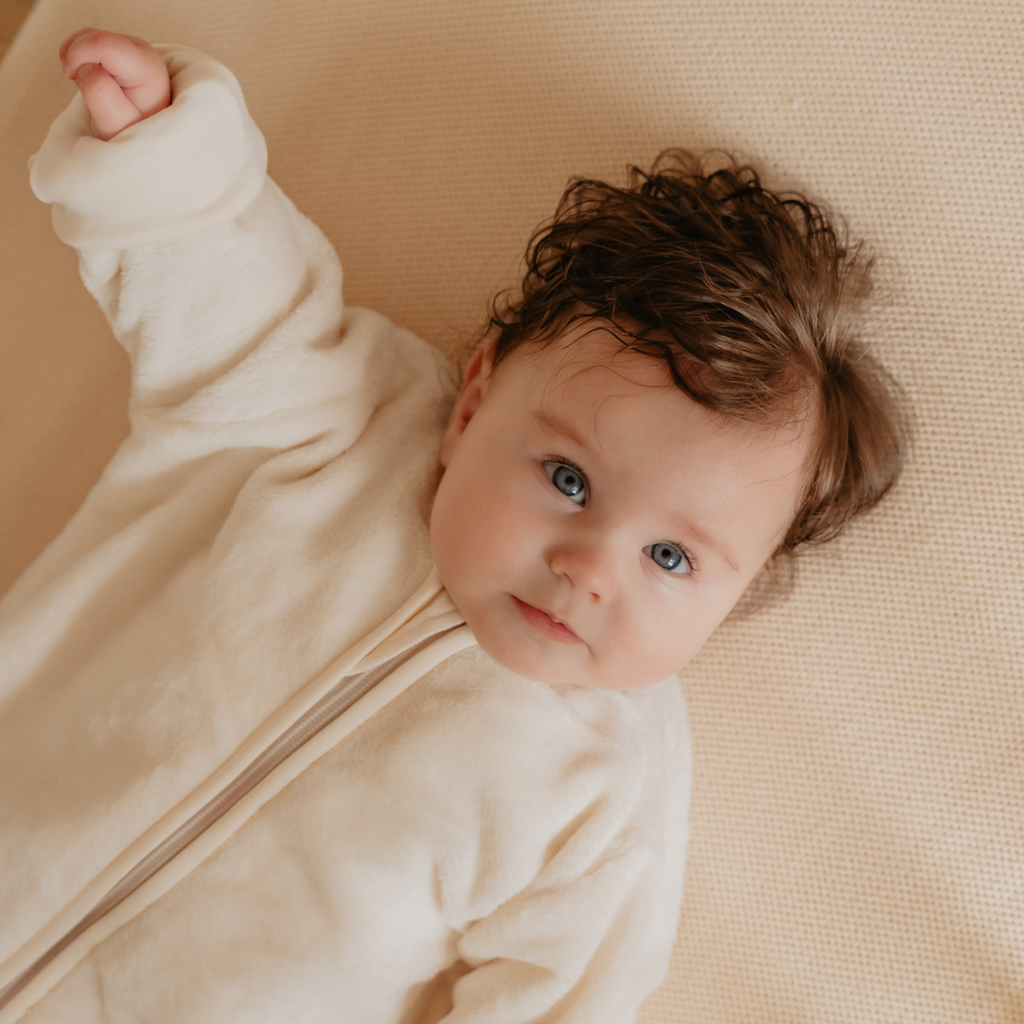 Baby met krullend haar in een Puckababy Bag Newborn Teddy Pure outfit ligt op een beige deken, heft één arm op en kijkt rustig omhoog.
