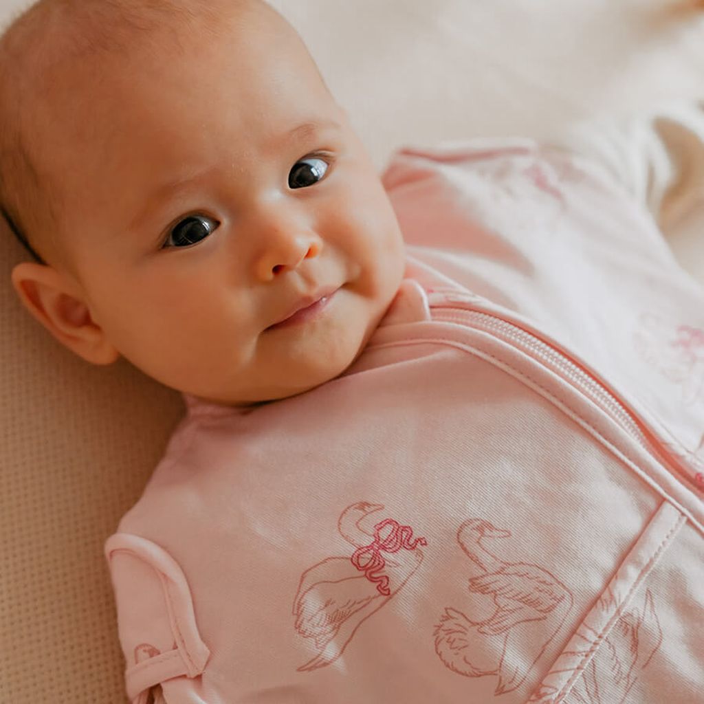 Een baby in een Puckababy Mini-slaapzak met Swans-print ligt op een zacht oppervlak en kijkt omhoog met een zachte uitdrukking.