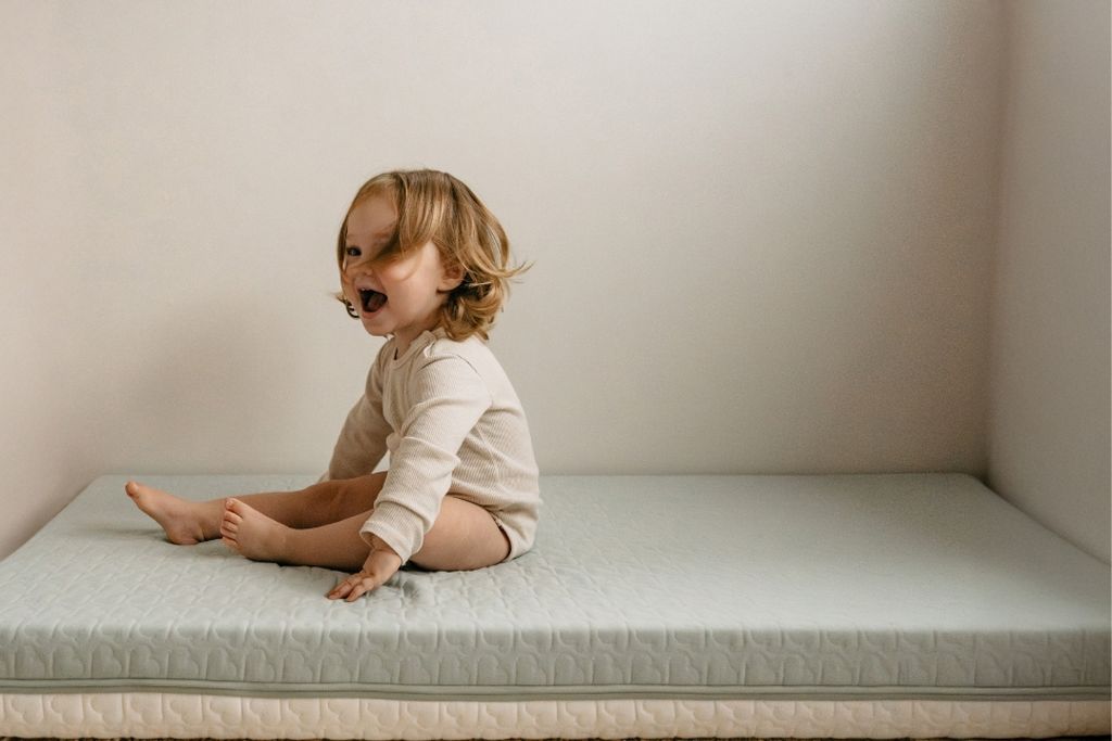 A toddler with long hair sits on a mattress in a corner, smiling and wearing a light-colored outfit.