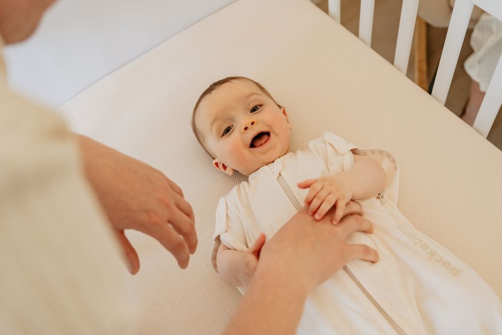 A baby lying in a crib, wearing a white outfit, smiling with an adult's hand touching their tummy.