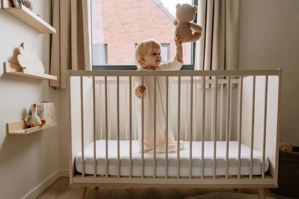 A child standing in a crib, holding a teddy bear, in a cozy room with shelves, toys, and a window in the background.