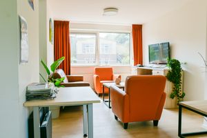 Lichte woonkamer met 3 tafels, 2 oranje stoelen, 1 bruine zitbank en tv met tv-meubel