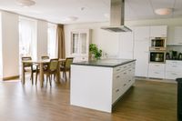 Een ruime, witte keuken met kookeiland en daarnaast een houten tafel met stoelen.